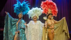 Imagem de três atrizes vestidas de vedetes, com roupas brilhosas e plumas na cabeça, no palco de um teatro