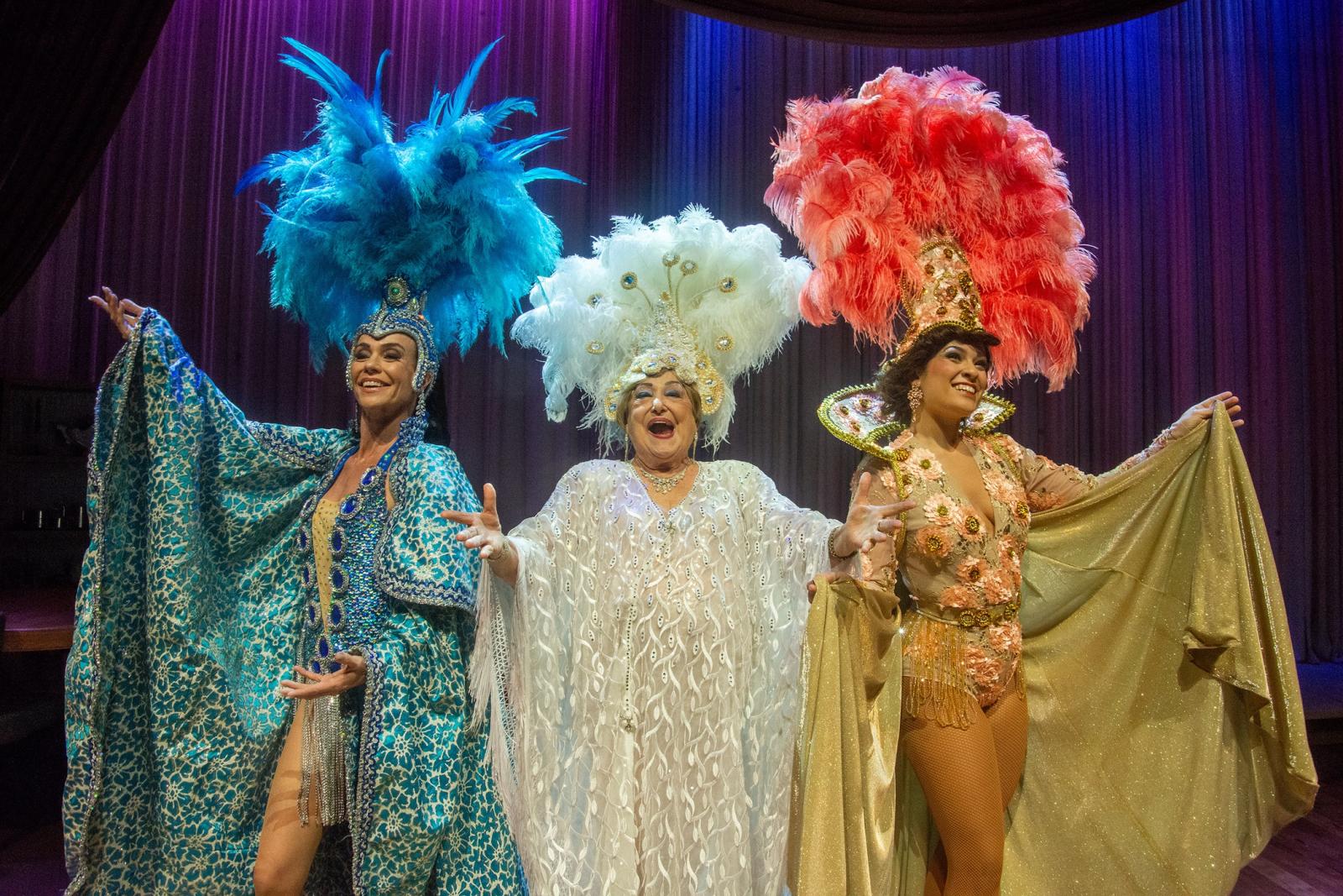 Imagem de três atrizes vestidas de vedetes, com roupas brilhosas e plumas na cabeça, no palco de um teatro