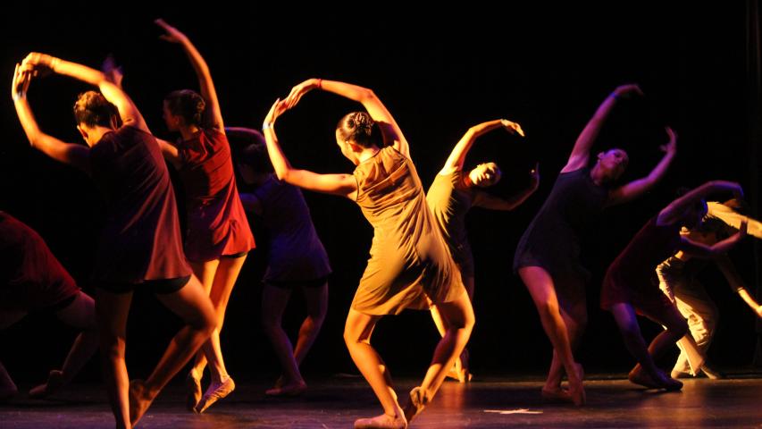 Grupo de bailarinas, vestidas cada uma de uma cor, fazem apresentação de dança contemporânea em um palco
