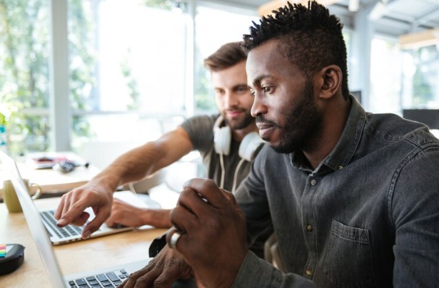 Foto de dois homens trabalhando diante de um notebook. Em primeiro plano, um homem negro. Em segundo plano, um homem branco.