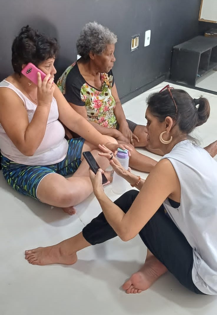 Fernanda Couzemenco (de branco) orienta mulheres sobre o uso do aplicativo. A imagem mostra três mulheres sentadas no chão, sendo que uma delas orienta as demais sobre o uso do aplicativo.
