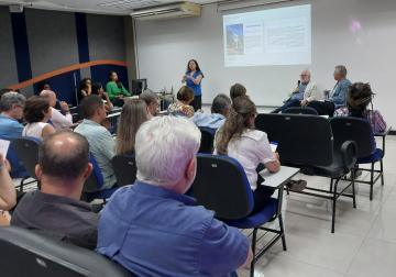 Foto das pessoas presentes no evento, de costas, sentadas nas cadeiras do auditório, com o reitor da Ufes à frente, sentado e falando ao microfone