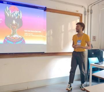 Foto do estudante Yuri Barreto de pé, usando camisa amarela e calça cinza, apresentando seu trabalho de conclusão de curso