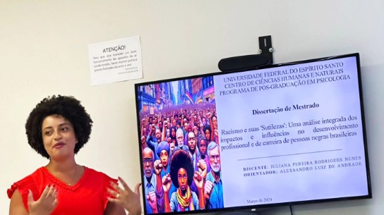 Foto da pesquisadora Juliana Nunes, em pé, apresentando sua pesquisa. Ela é uma mulher negra, de cabelos cursos e veste vestido vermelho.