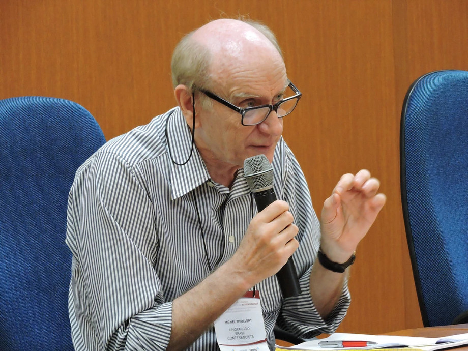 Foto do professor Michel Thiollent em uma palestra, falando ao microfone.