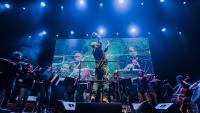 Foto de uma apresentação da orquestra onde aparecem o maestro e os músicos em um ambiente com luz azul