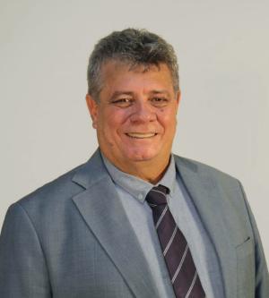 Fotografia de homem de pele clara e cabelos grisalhos, sorrindo e usando terno cinza, camisa azul clara e gravada azul escura com listas em diagonal