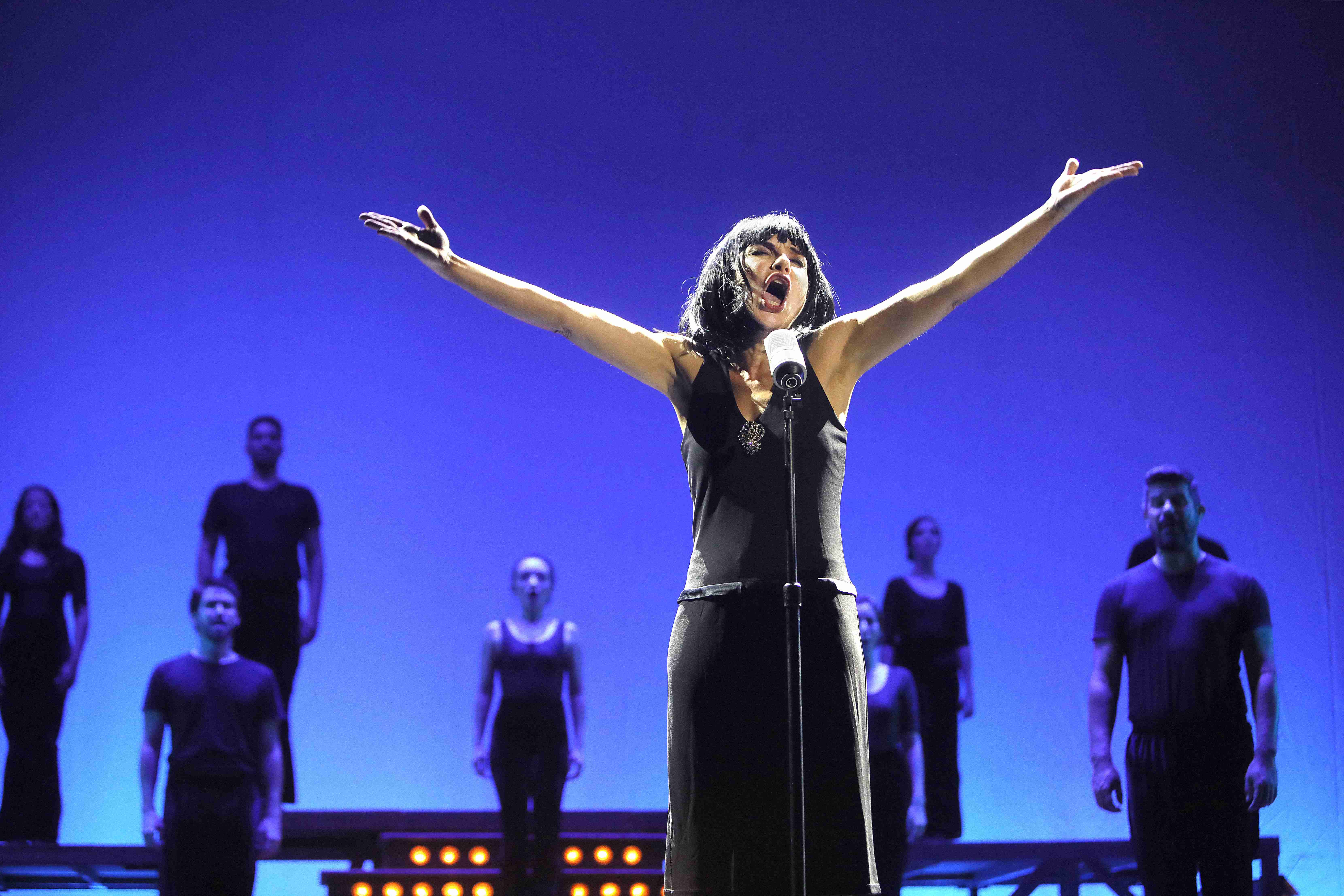 Cena do espetáculo "Bibi, uma vida em musical" Foto de uma cena do espetáculo que mostra a atriz cantando de frente para um microfone, com os braços abertos para o alto. Atrás, bailarinos com roupas pretas fazem coro.