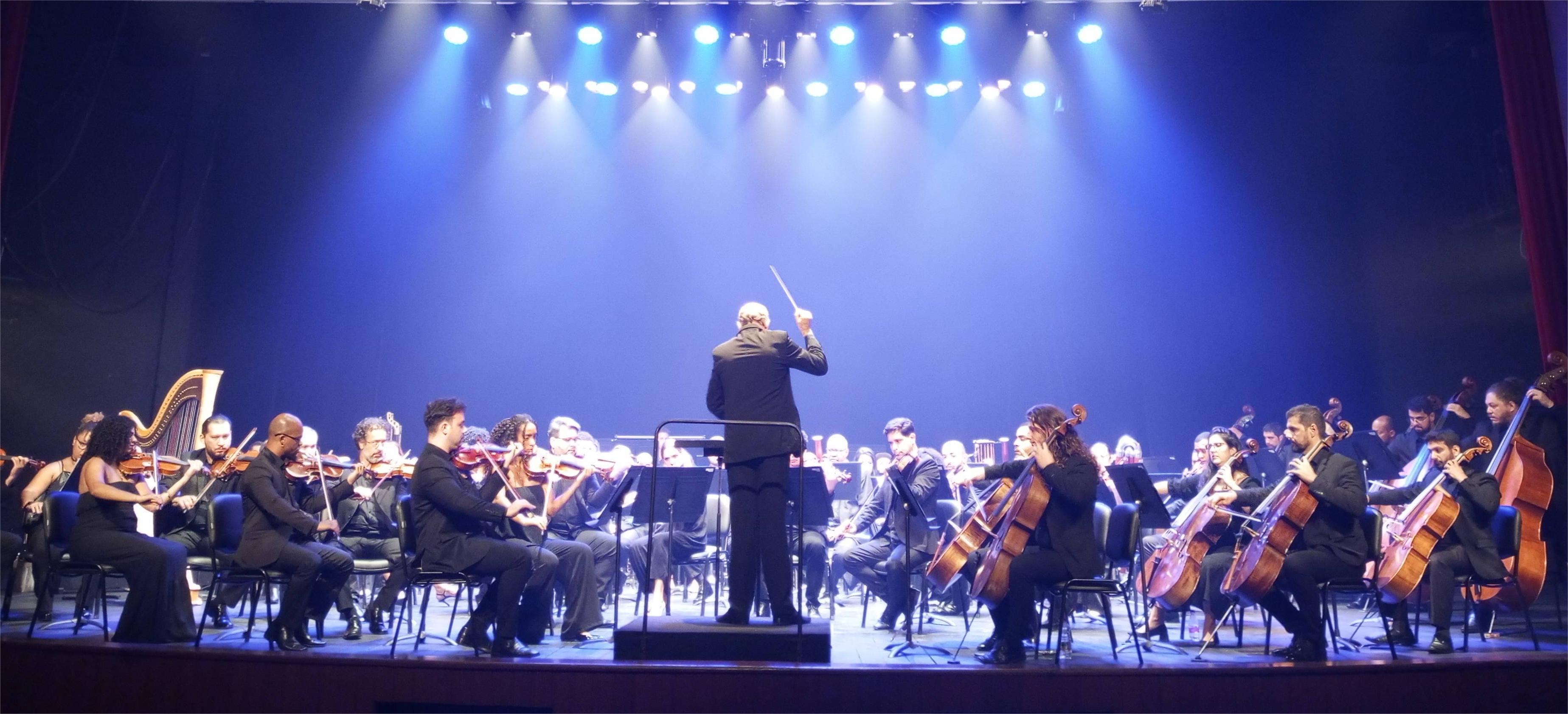 Foto da orquestra sinfônica com o maestro de costas regendo os músicos a sua frente