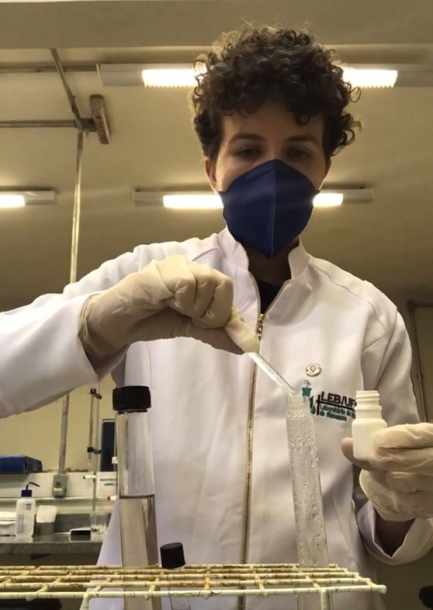 A pesquisa tem o apoio do Laboratório Multiusuário de Energia da Biomassa da Ufes Pesquisadora com jaleco e máscara em um laboratório colocando alguma substância dentro de um tubo de ensaio