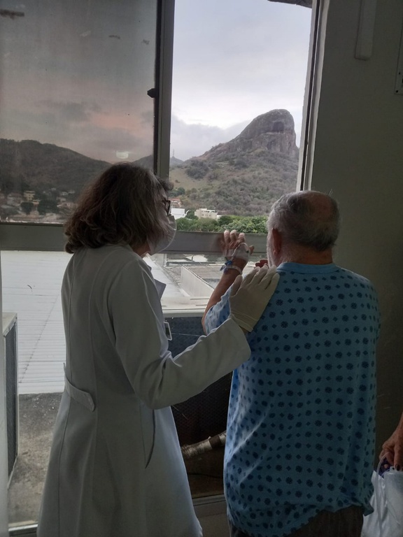 Integrante da equipe de cuidados paliativos do Hucam cuidando de um paciente Foto que mostra um integrante da equipe de cuidados paliativos do Hucam cuidando de um paciente. Ambos estão em pé, de frente para uma janela
