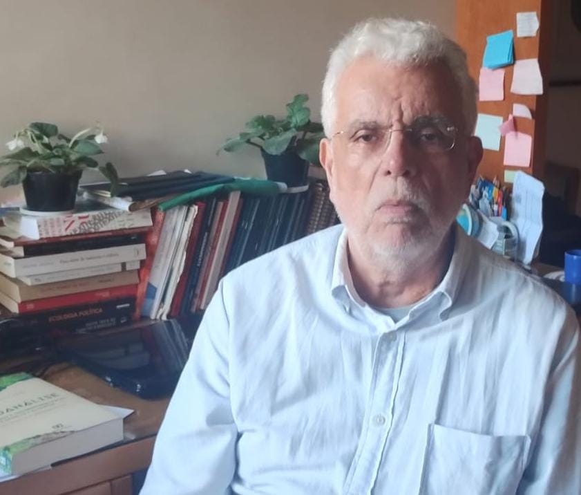 Professor Roberto Simões Foto do professor Roberto Simões sentado, vestindo uma camisa branca, ao lado de uma mesa com alguns livros