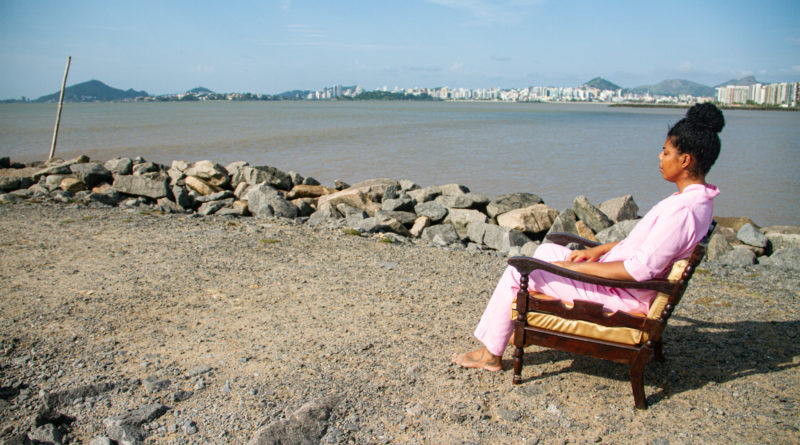 Rubiane Maia em uma cena do filme Presença Foto da artista Rubiane Maia sentada em uma poltrona em um píer de frente para o mar, em uma cena do filme Presença