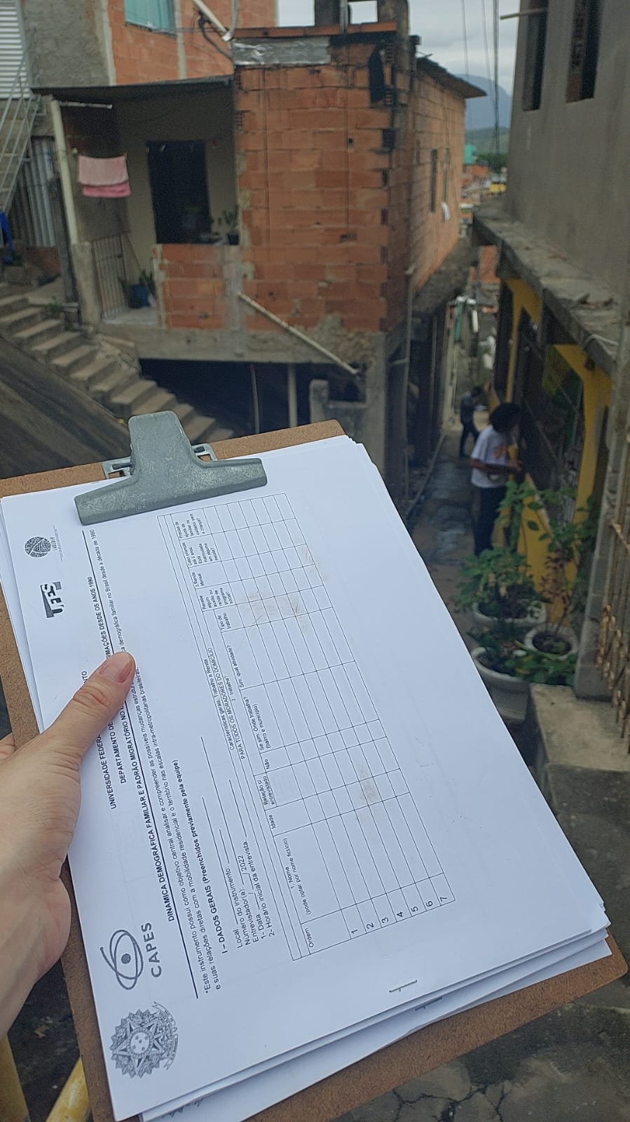 Foto de uma mão segurando uma prancheta com formulários de pesquisa. Ao fundo, casas de tijolos