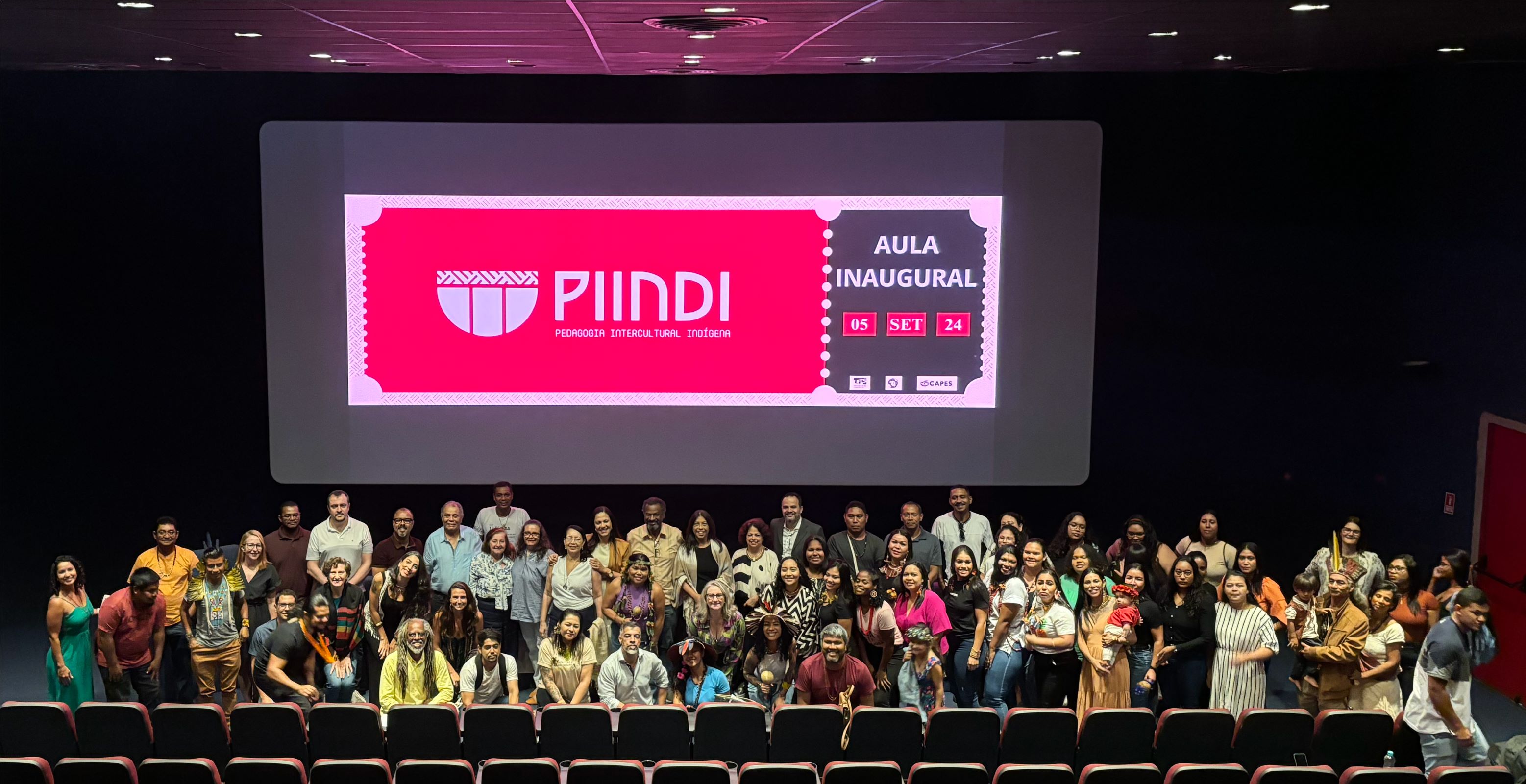 Abertura da aula inaugural do PIINDI Foto da abertura da aula inaugural, com todos os presentes posando para a foto. Atrás, projetada na tela do cinema, a logomarca do curso e a data da aula.
