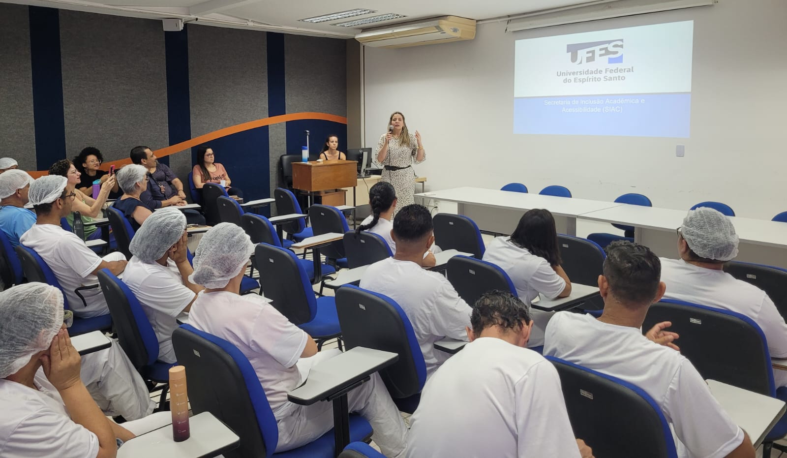 A secretária de Inclusão Acadêmica e Acessibilidade, Cinthya Oliveira, na formação para os trabalhadores. Foto da secretária Cinthya Oliveira falando para os trabalhadores no auditório