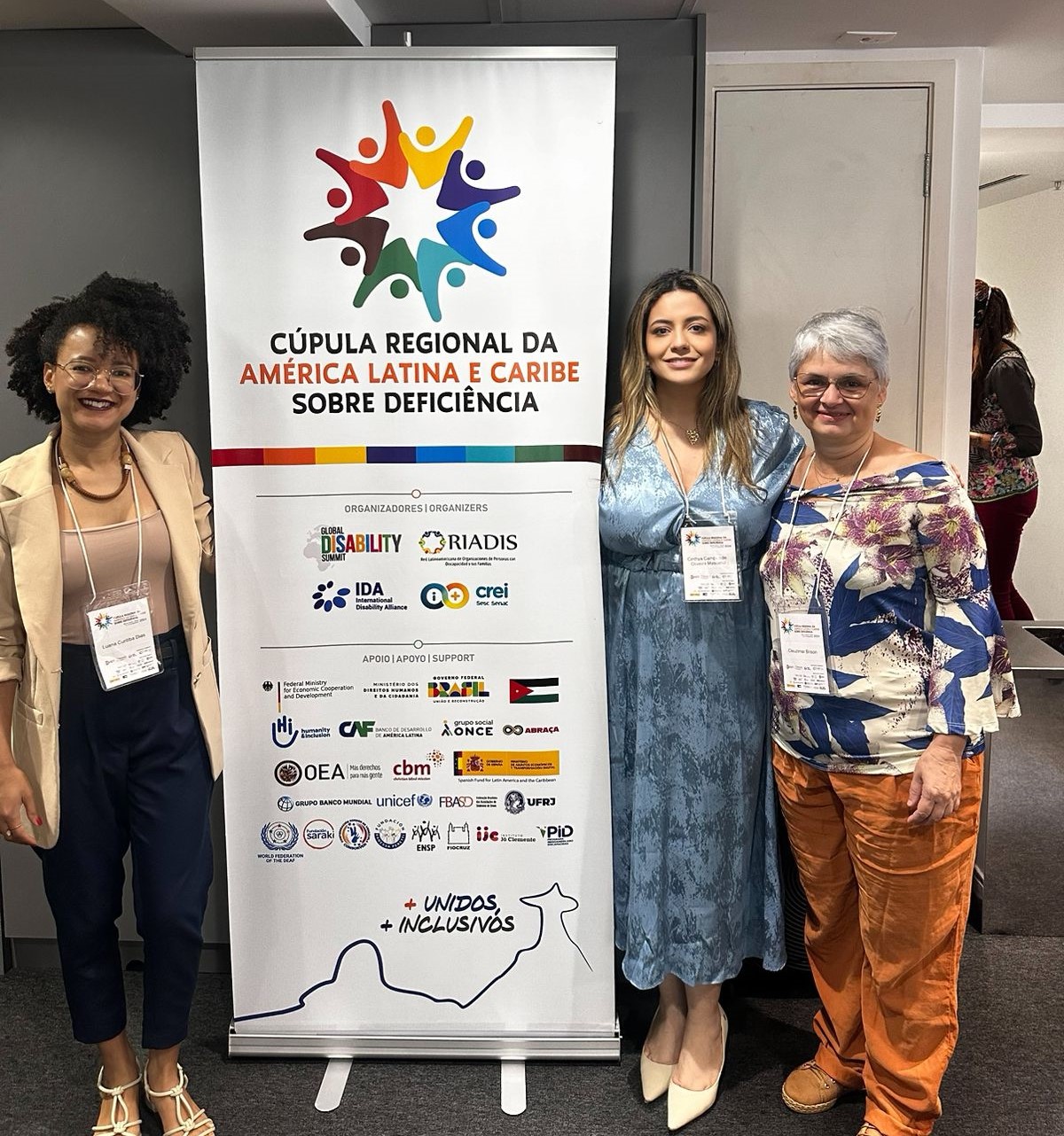 A assistente social Luana Dias, a secretária Cinthya de Oliveira e a pedagoga Cleuzimar Brizon Foto da assistente social Luana Dias, da secretária Cinthya de Oliveira, e da pedagoga Cleuzimar Brizon ao lado de um banner do evento
