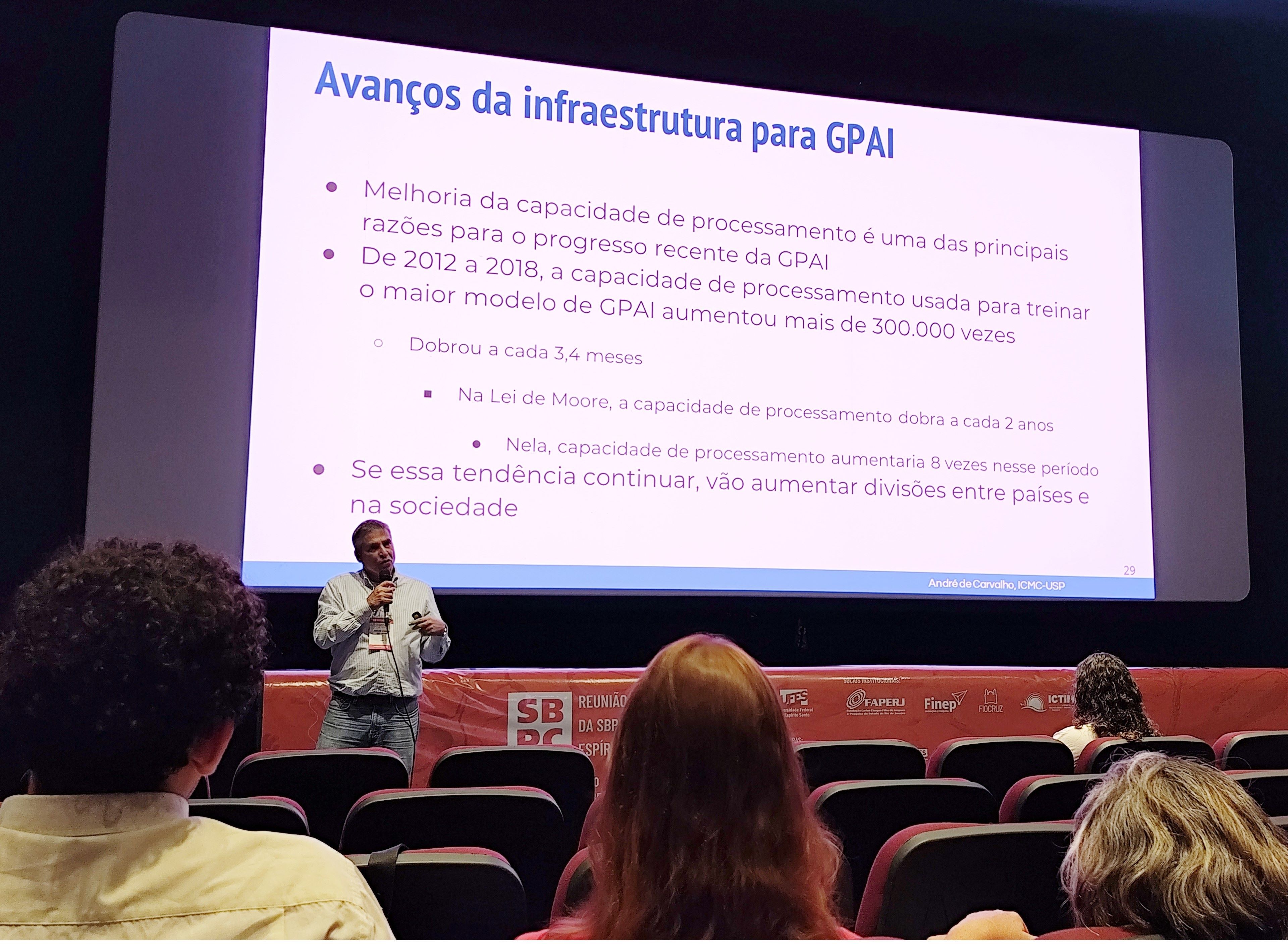 O professor André Carlos de Carvalho Foto do professor no palco do Teatro, em frente a um slide
