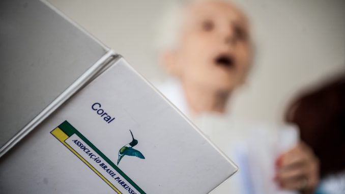 Foto de uma senhora cantando, tendo nas mãos um livro do coral da Associação Brasil Parkinson