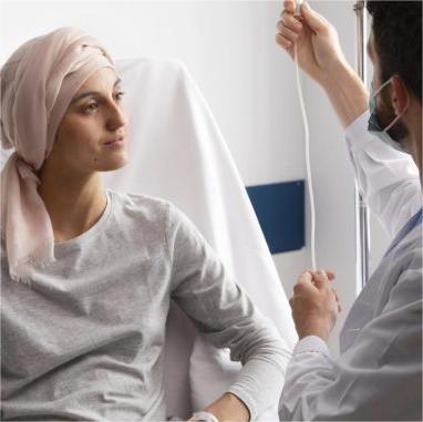 Foto de paciente sentada em leito hospitalar, utilizando lenço bege na cabeça e sendo atendida por enfermeiro