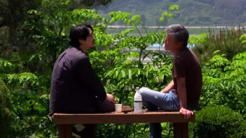 Cena de O que a natureza te conta, onde aparecem dois homens sentados em um banco, conversando