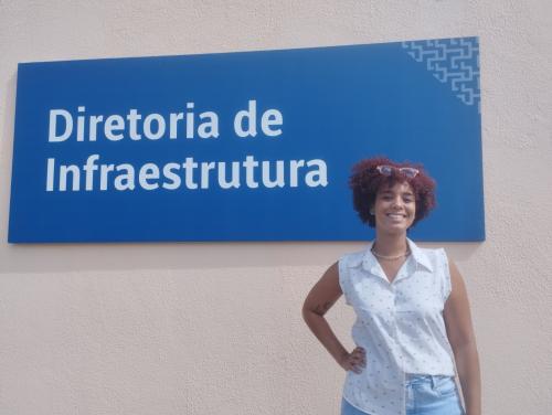 Foto de Marília Santana em frente à placa da Diretoria de Infraestrutura