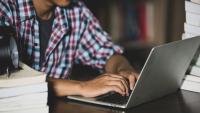 Foto de um jovem digitando em um notebook sobre uma mesa com livros