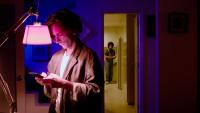 Cena de Labirinto dos Garotos Perdidos em que aparece um homem digitando no celular em uma sala com baixa iluminação enquanto um jovem observa de longe