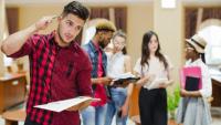 Imagem de um estudante em pé, segurando um caderno e coçando a cabeça em sinal de dúvida. Ao fundo, outros estudantes conversam entre si