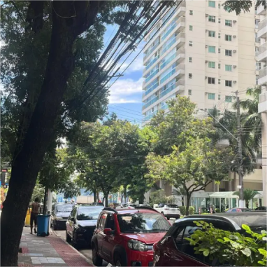 Foto de carros estacionados numa rua, com árvore na calçado ao lado esquerdo e prédio com apartamentos ao lado direito