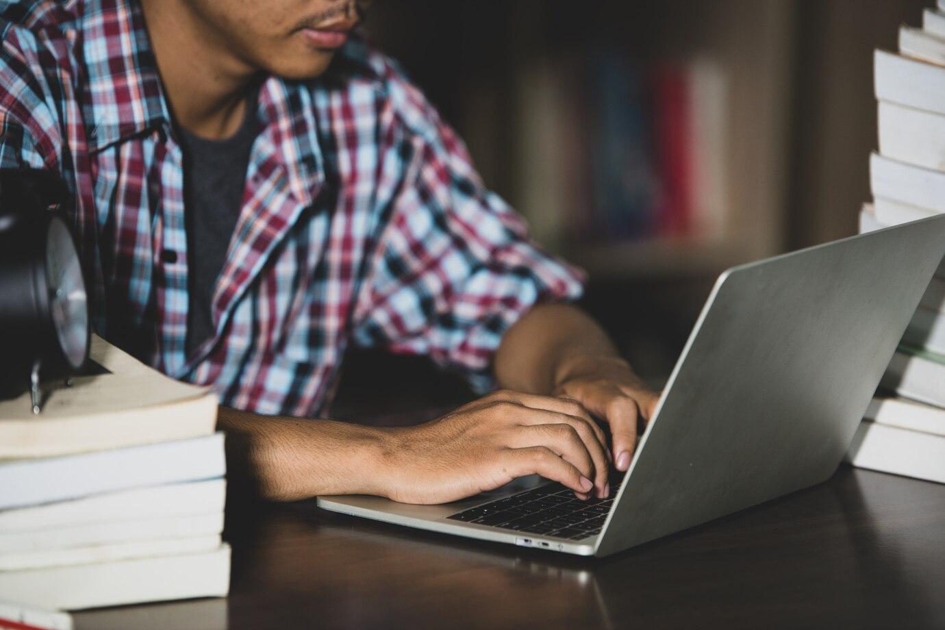 Foto de um jovem digitando em um notebook sobre uma mesa com livros