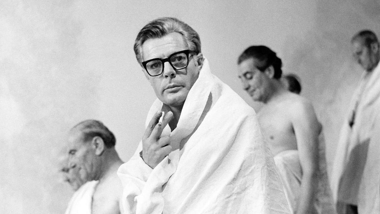 Cena do filme em que o protagonista aparece enrolado em um pano branco fumando um cigarro. Atrás dele, outros homens passam, também enrolados em um tecido branco