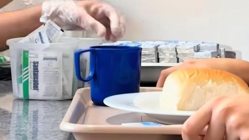 Foto de mãos segurando uma bandeja onde há um prato com um pão e uma xícara. Na imagem aparecem ainda duas mãos colocando um sachê de açúcar sobre a bandeja